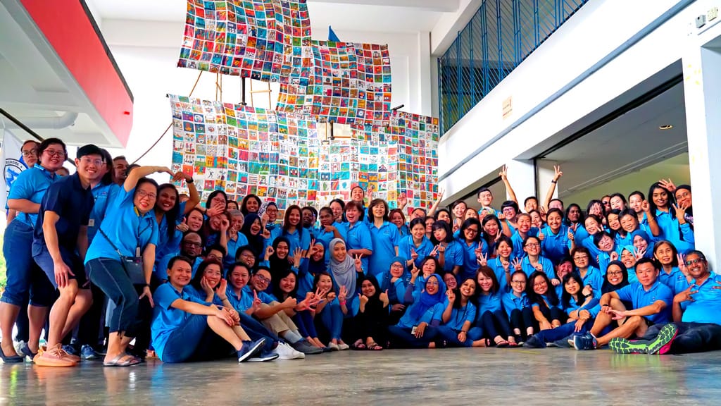Large group of people in blue shirts posing indoors in front of a colorful patchwork sail ship display.